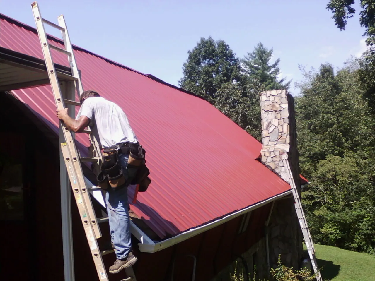 Roofer on a ladder beside a red metal roof and stone chimney — Soffit & Fascia Repair by Roofing Brothers in Annville
